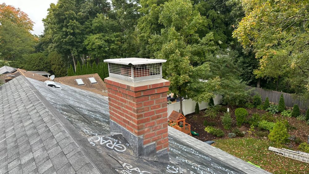 A brick chimney on a gray shingled roof with a metal chimney cap, surrounded by green trees.