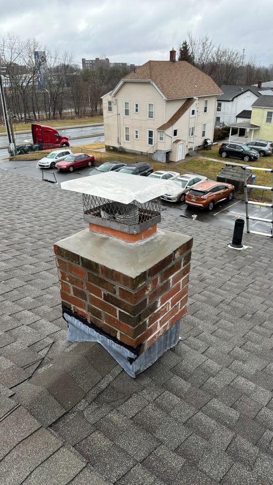 Brick chimney with a metal cap on a shingle roof, cars and buildings in the background on a cloudy day.