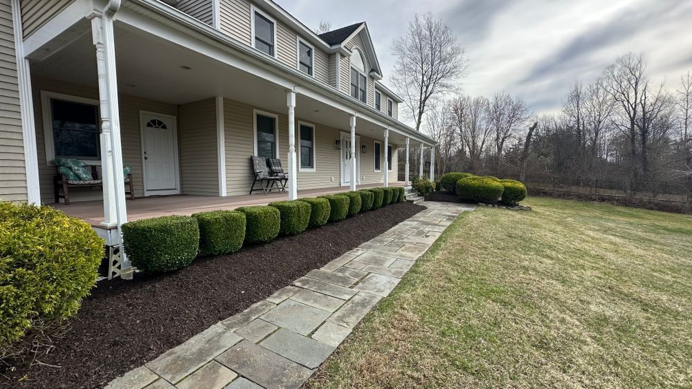 A two-story beige house with a wraparound porch, pathway, and landscaped shrubs along the walkway on a cloudy day.