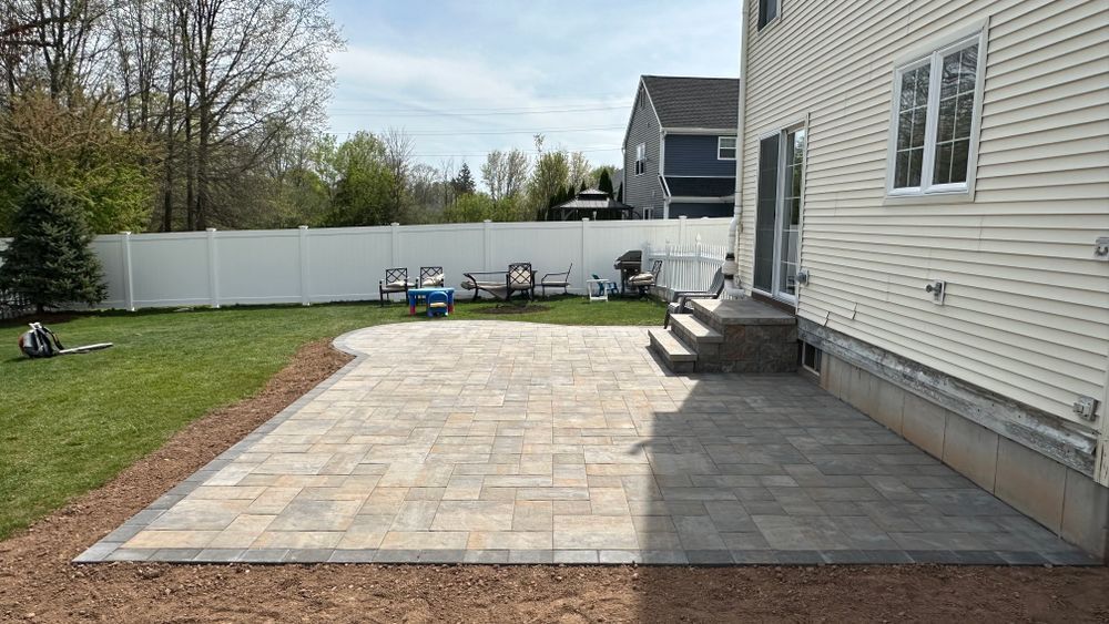A backyard patio paved with light-colored bricks next to a beige house. A white fence and grass surround the patio.