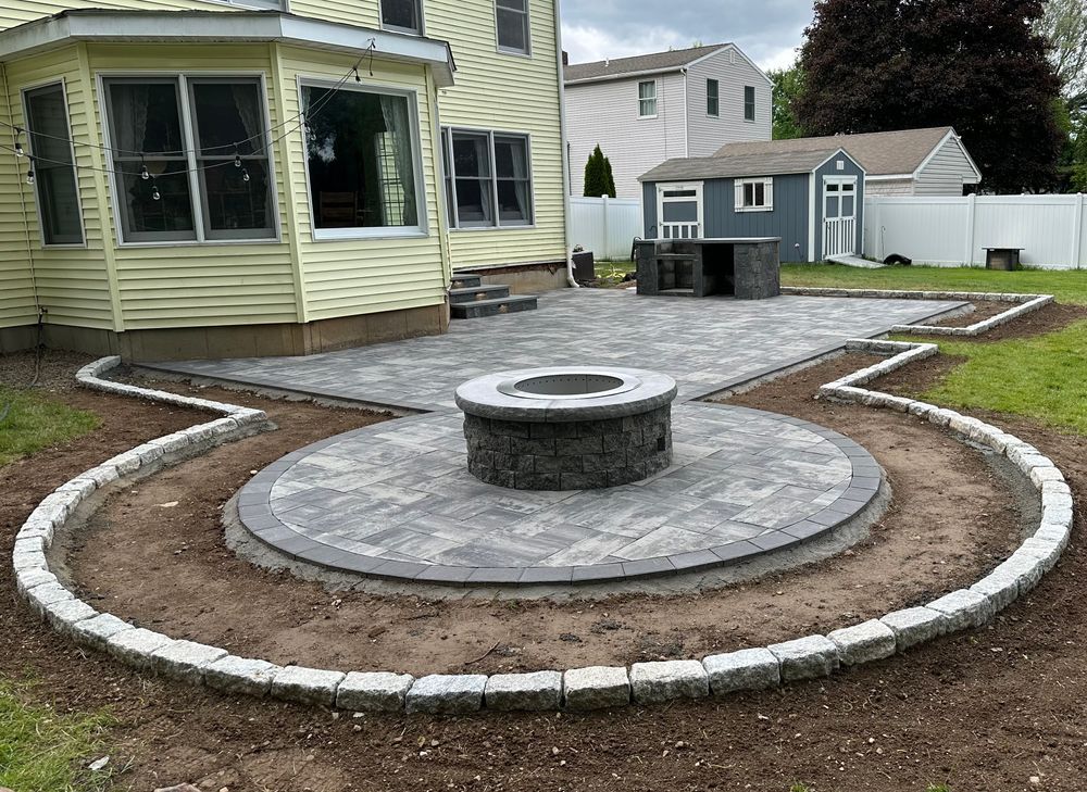 Backyard patio with a circular fire pit, surrounded by pavers and a border, near a yellow house.