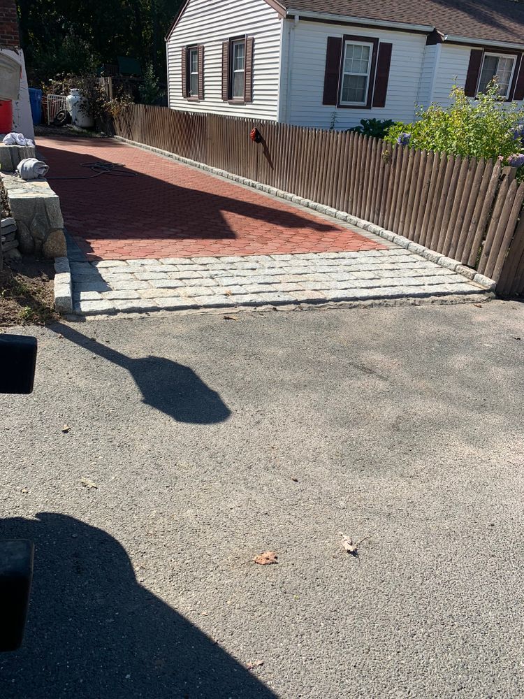 Asphalt driveway leads to a paved brick entry area and wooden fence bordering a white house.