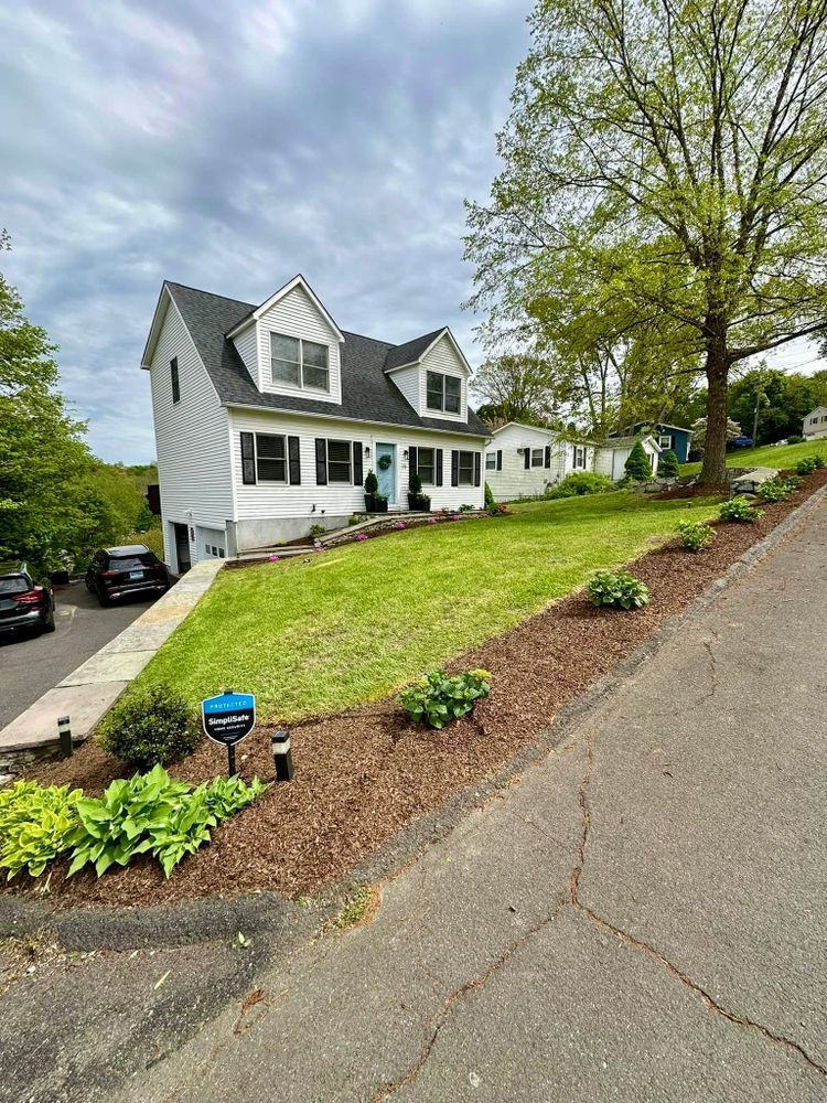 White two-story house with dormers on a grassy hill. A blue front door and landscaping with a cloudy sky.