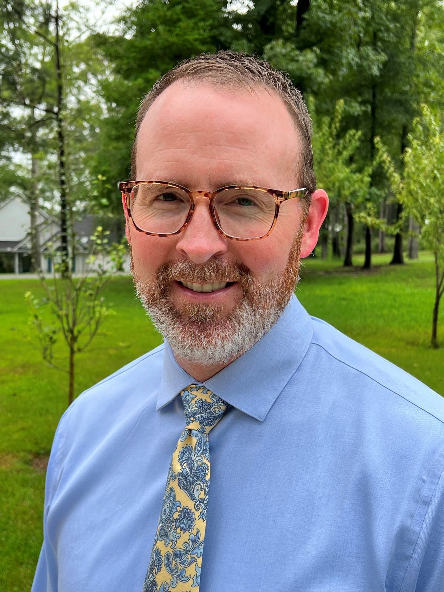 Smiling man with glasses and beard in a blue shirt and patterned tie standing outdoors in a green park.