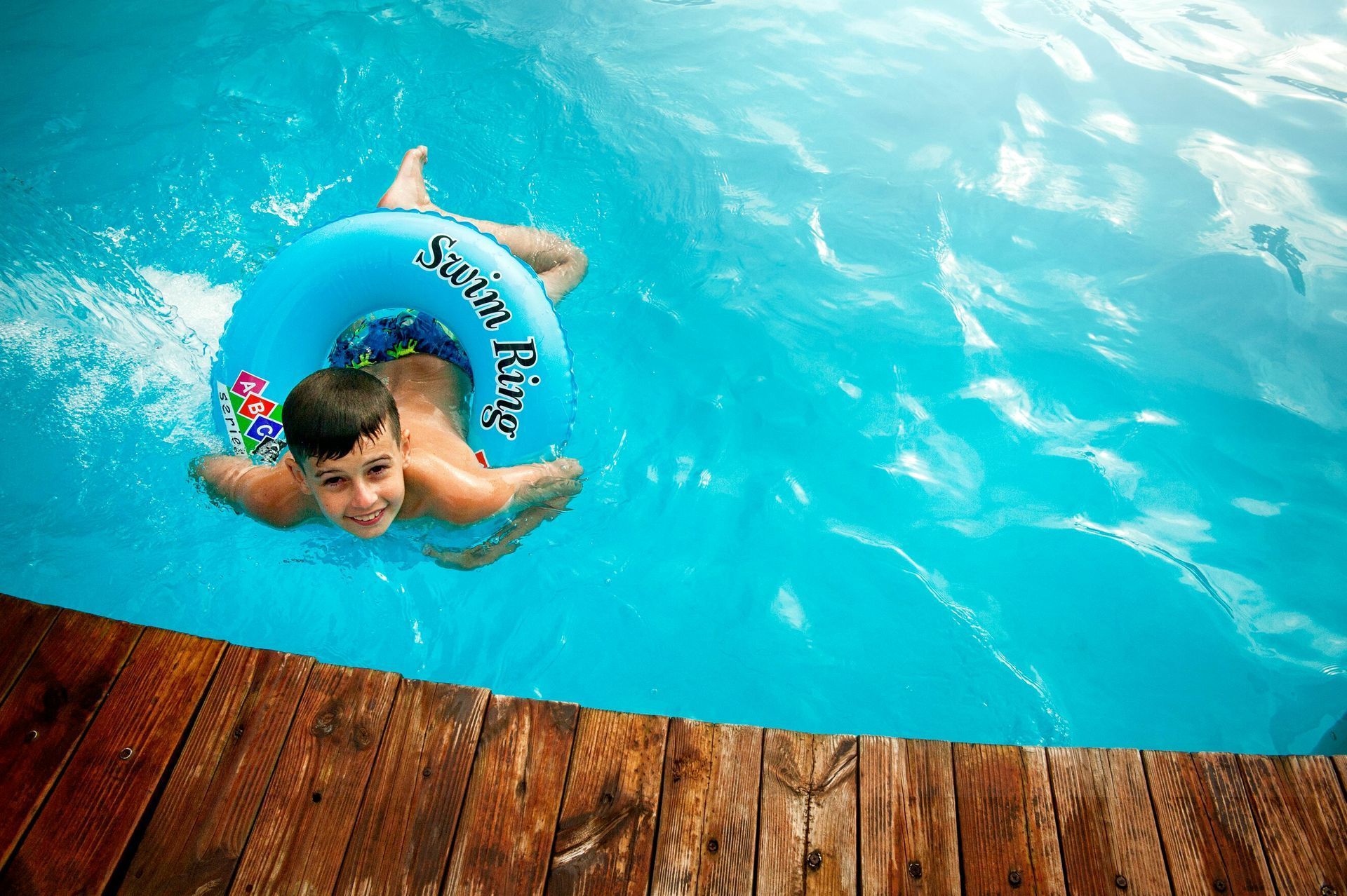 A child smiles while swimming in a bright blue pool using a blue inflatable ring next to a wooden deck.