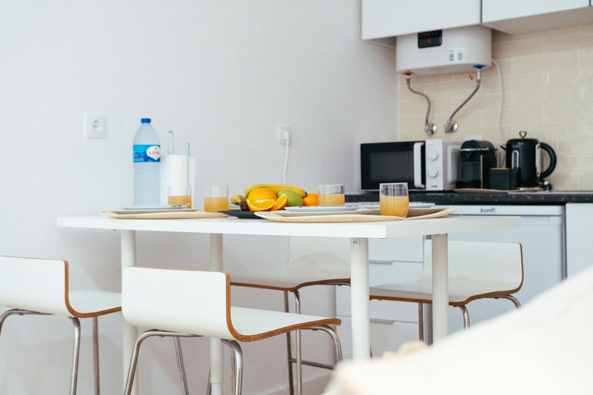 White dining table set with four chairs, with fruit and drinks, and microwave in a small kitchen.