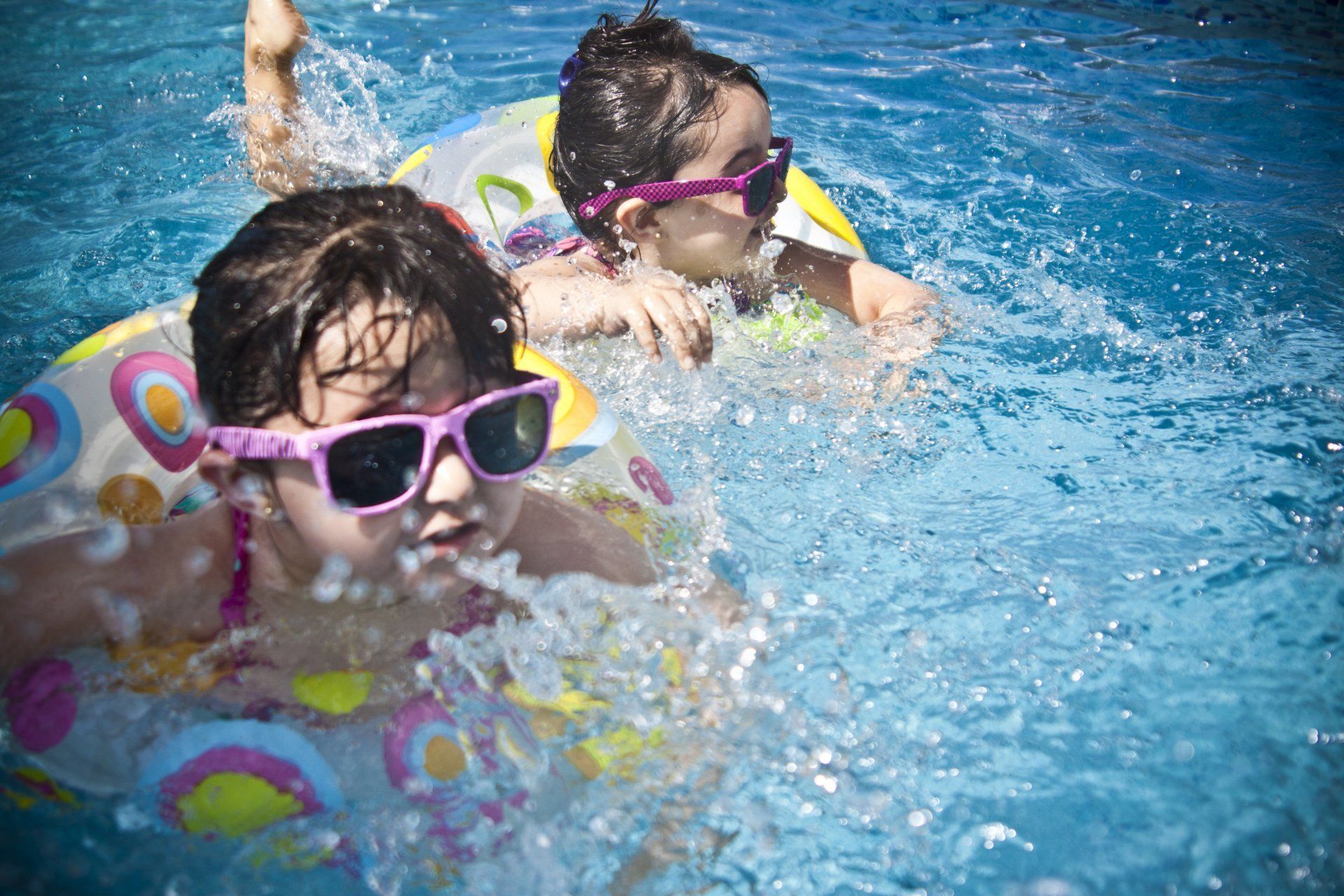 Two people wearing pink-rimmed sunglasses swimming in a pool with patterned inflatable swim rings.