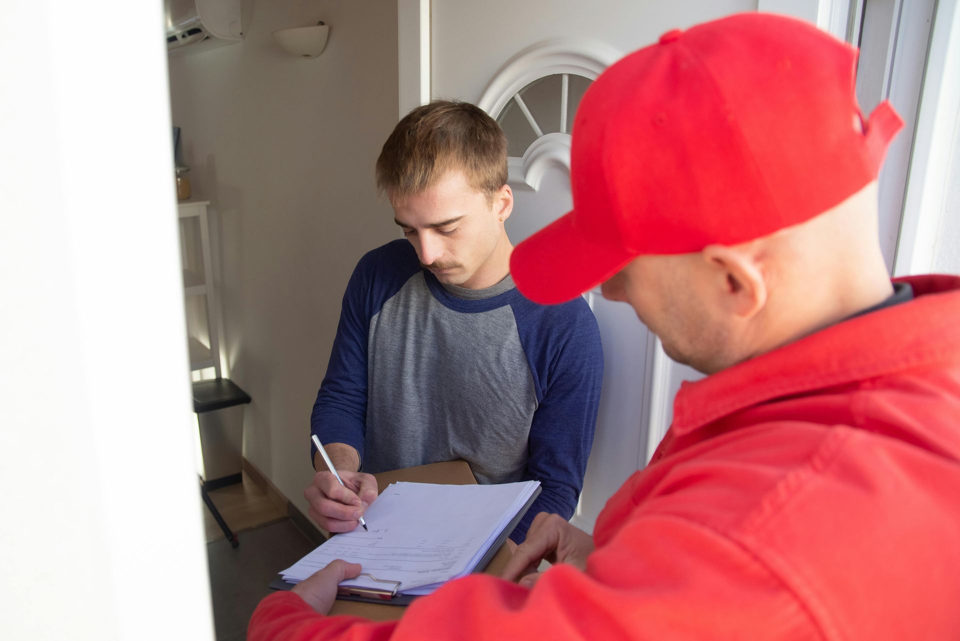 Delivery person in red cap/jacket hands clipboard to person signing for package at doorway.