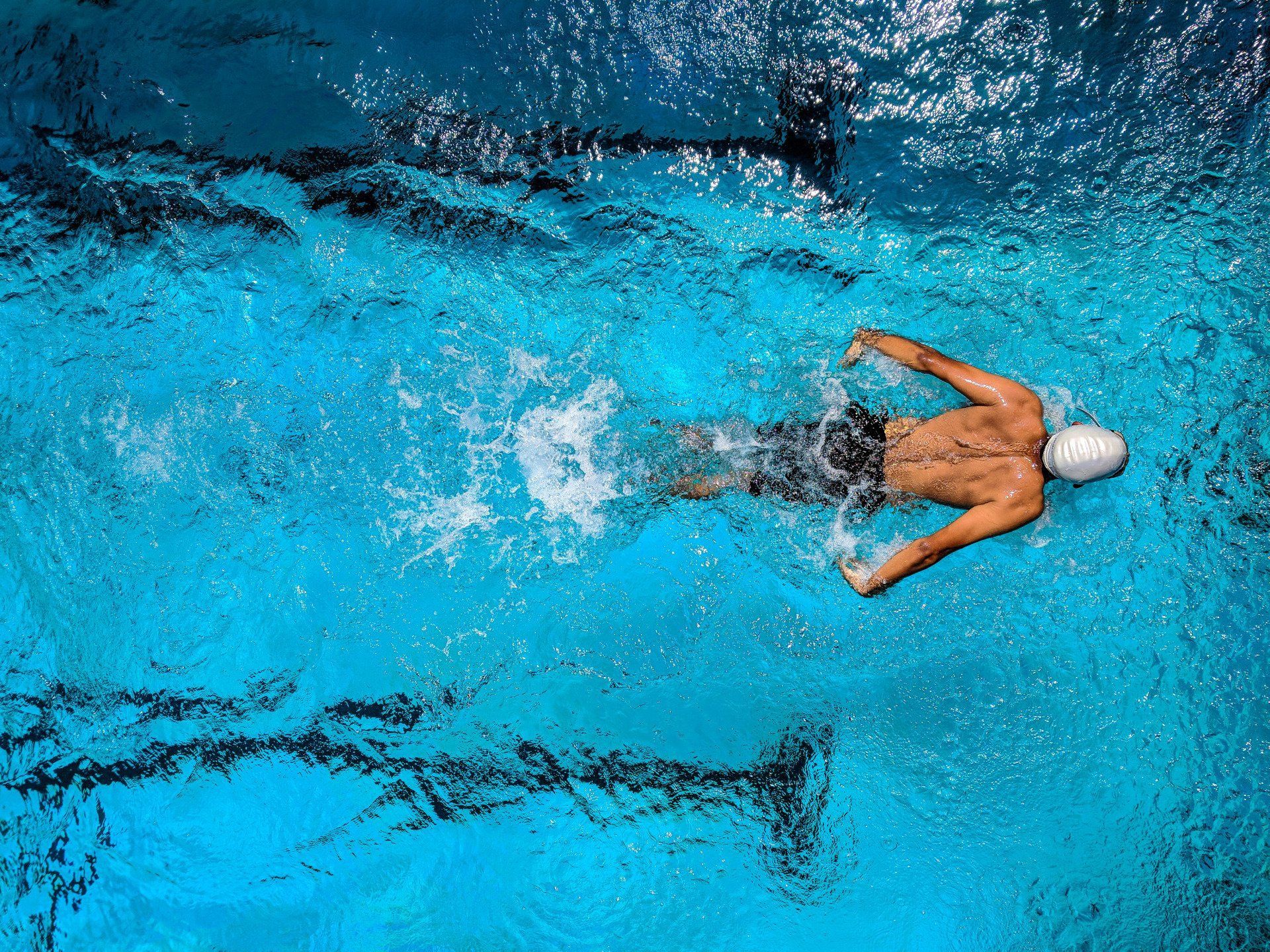 A swimmer with a white cap performing the butterfly stroke in a bright blue pool, viewed from directly above.