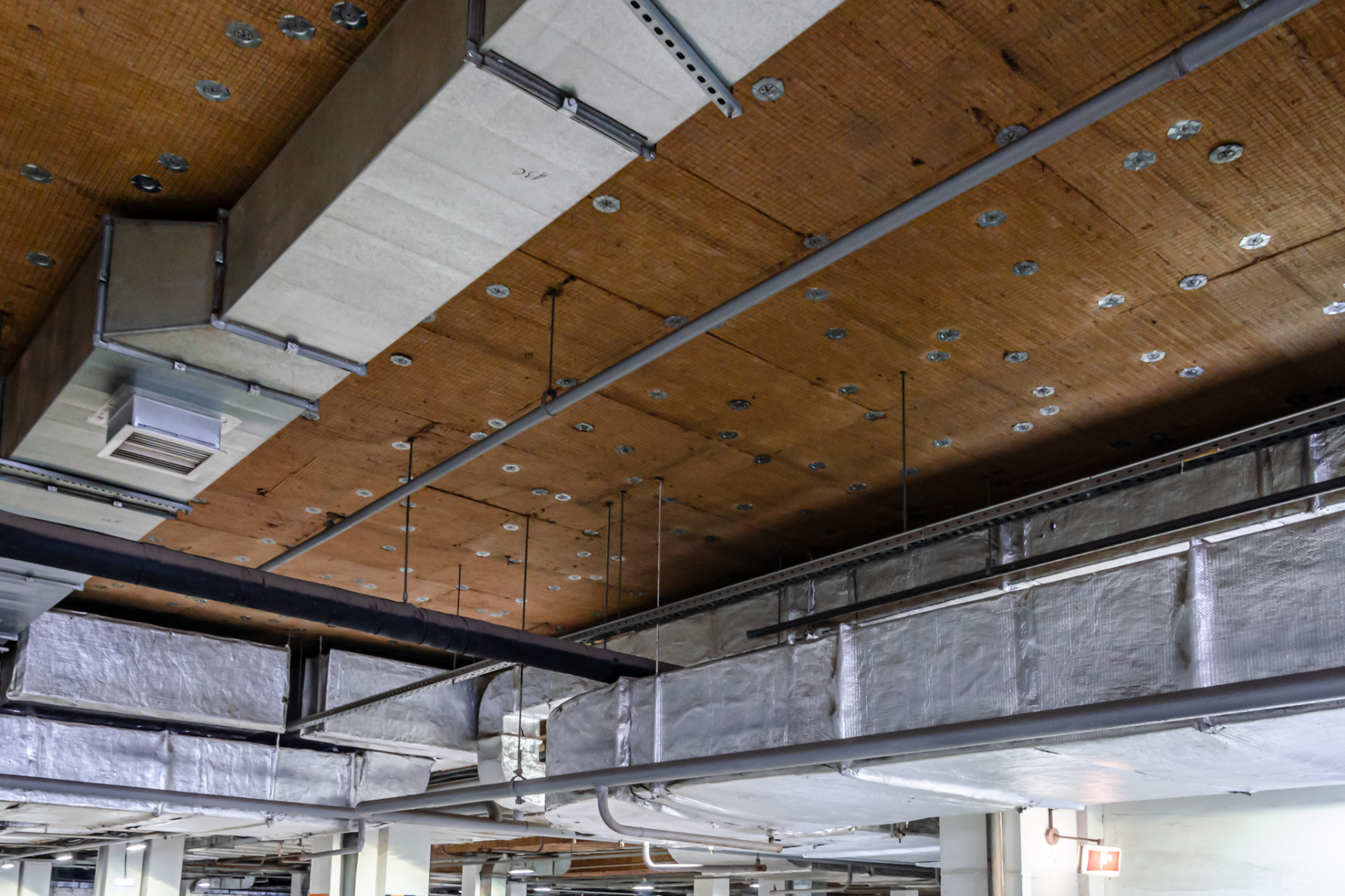 Exposed ceiling in a building with wooden panels, large silver air ducts, and industrial pipes and fixtures.