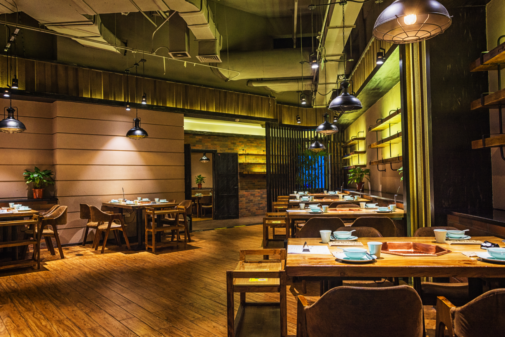 A warmly lit restaurant interior with wooden tables and chairs, pendant lights, and shelves on beige-toned walls.