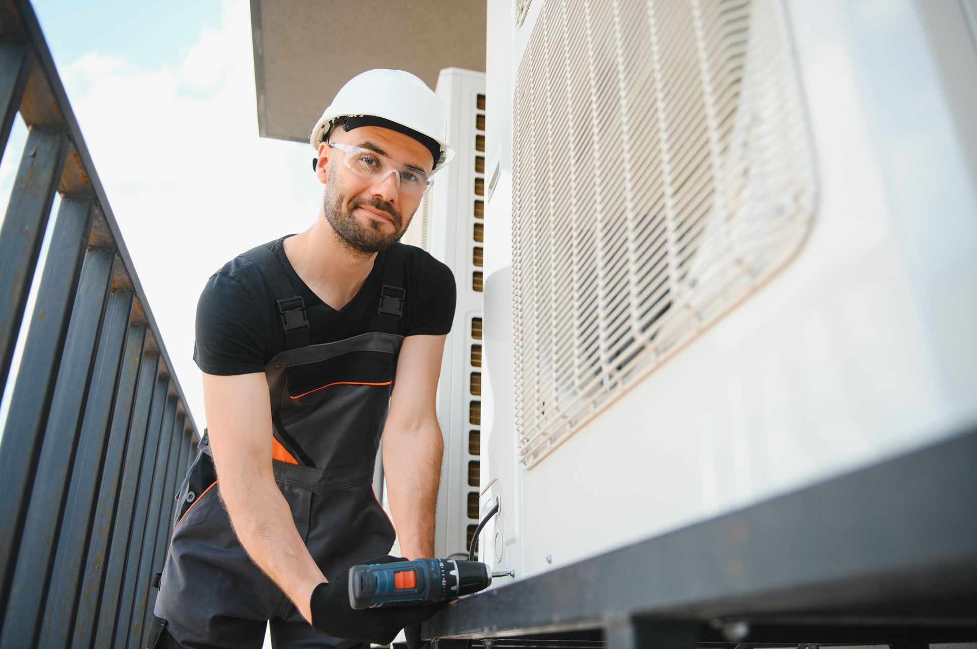A technician in a hard hat and safety glasses uses a drill while servicing an outdoor air conditioning unit.