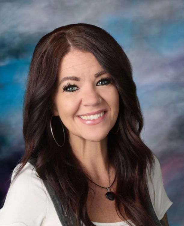 A woman wearing a yellow cardigan and a white shirt is smiling in front of a blue background.