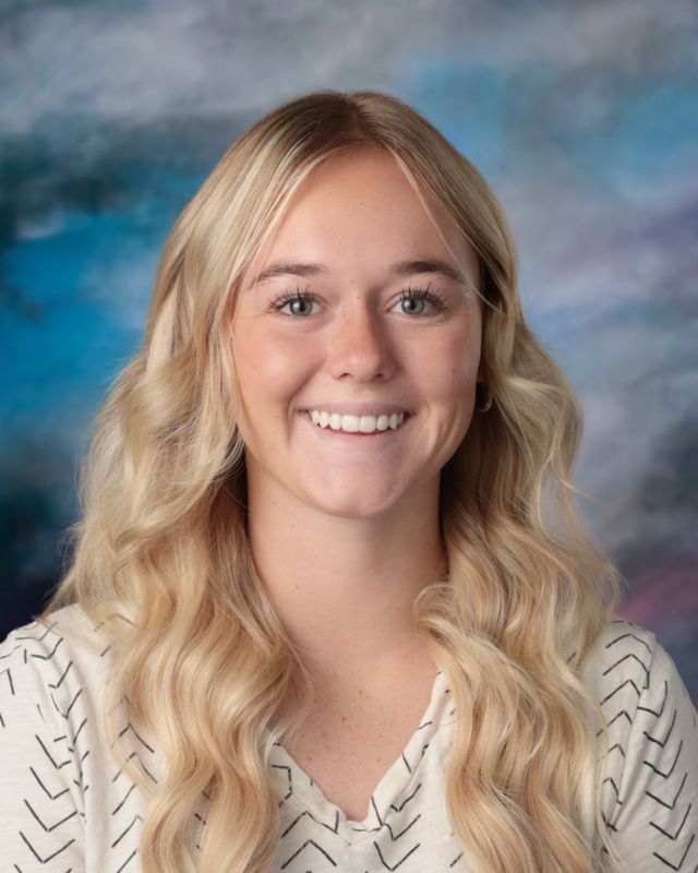 A young woman with long blonde hair is smiling for a picture in front of a blue background.