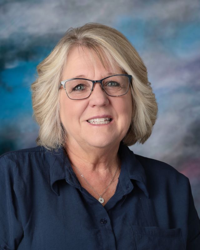 A woman wearing glasses and a gray shirt is smiling for the camera.