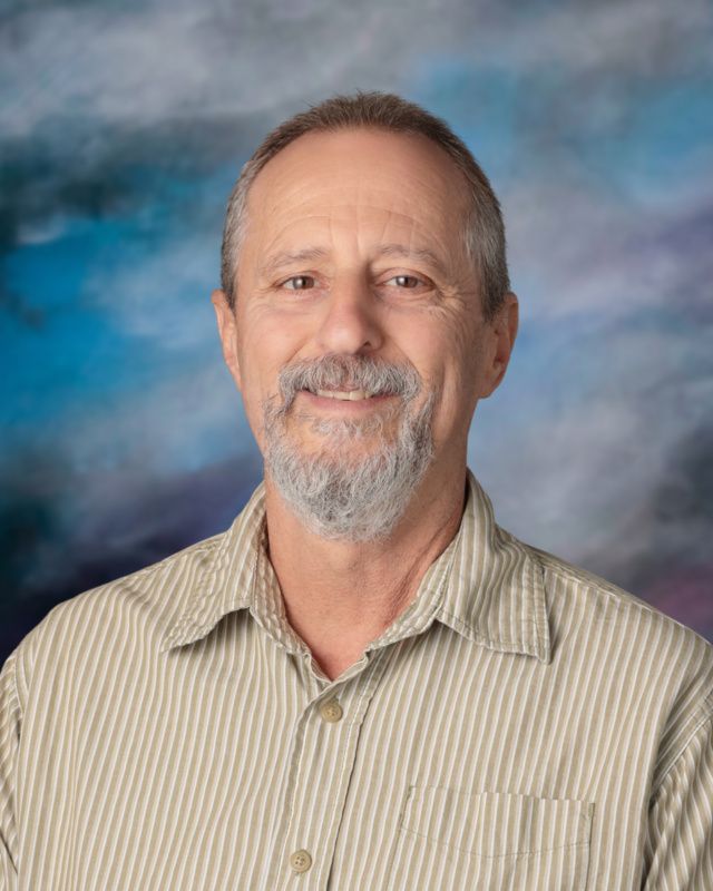 A man with glasses and a beard is standing in front of a blue background.