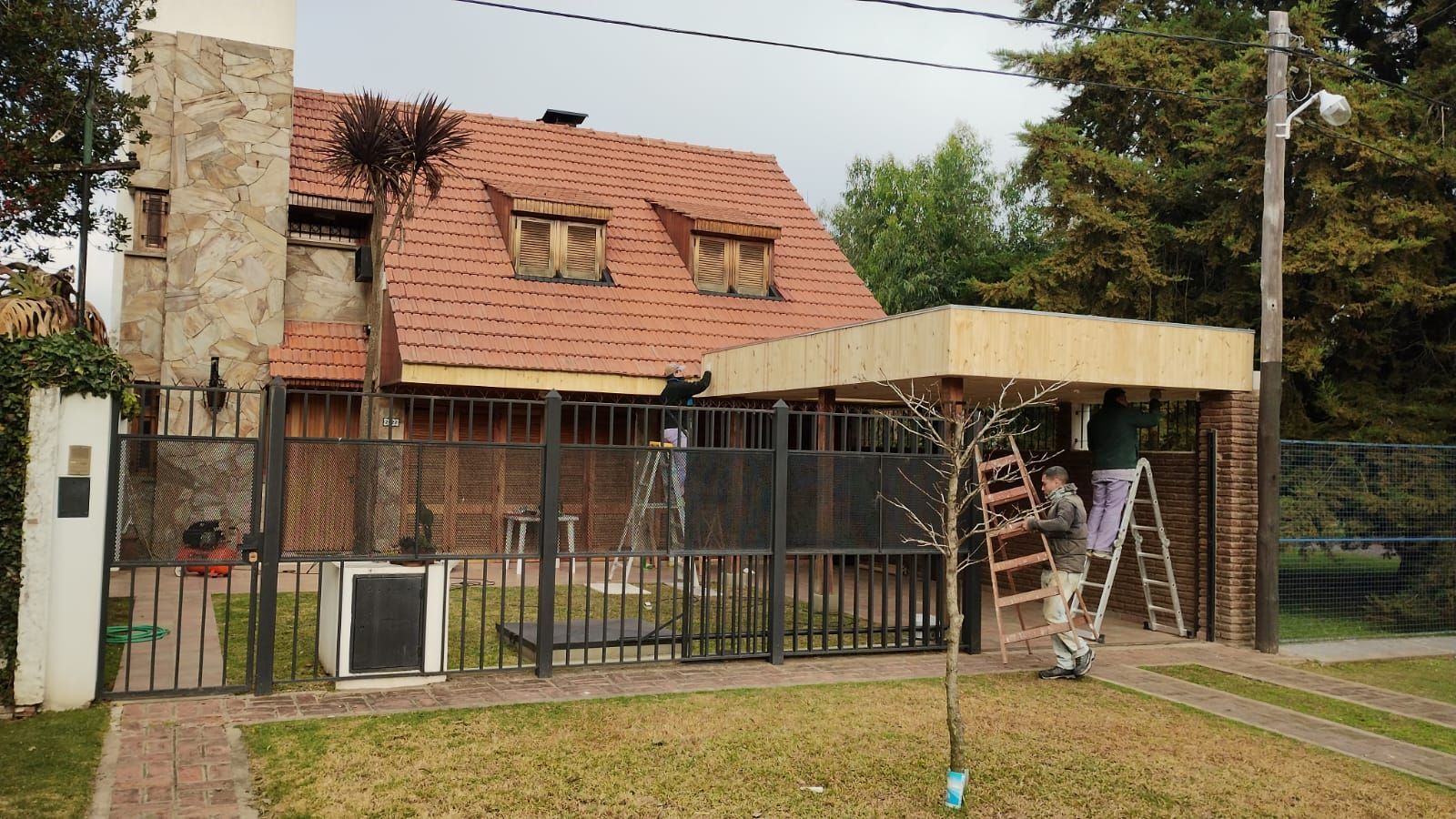 Casa con techo y portón rojos, con una escalera bajo un toldo beige en un patio con césped.