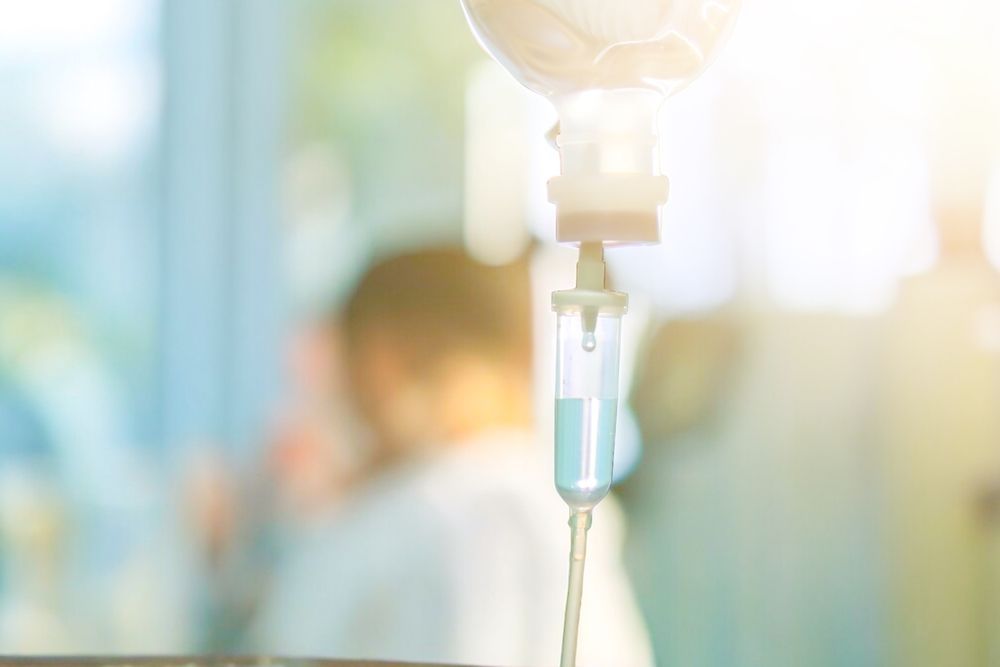 IV drip bag with fluid, blurred figure in background, bright light.