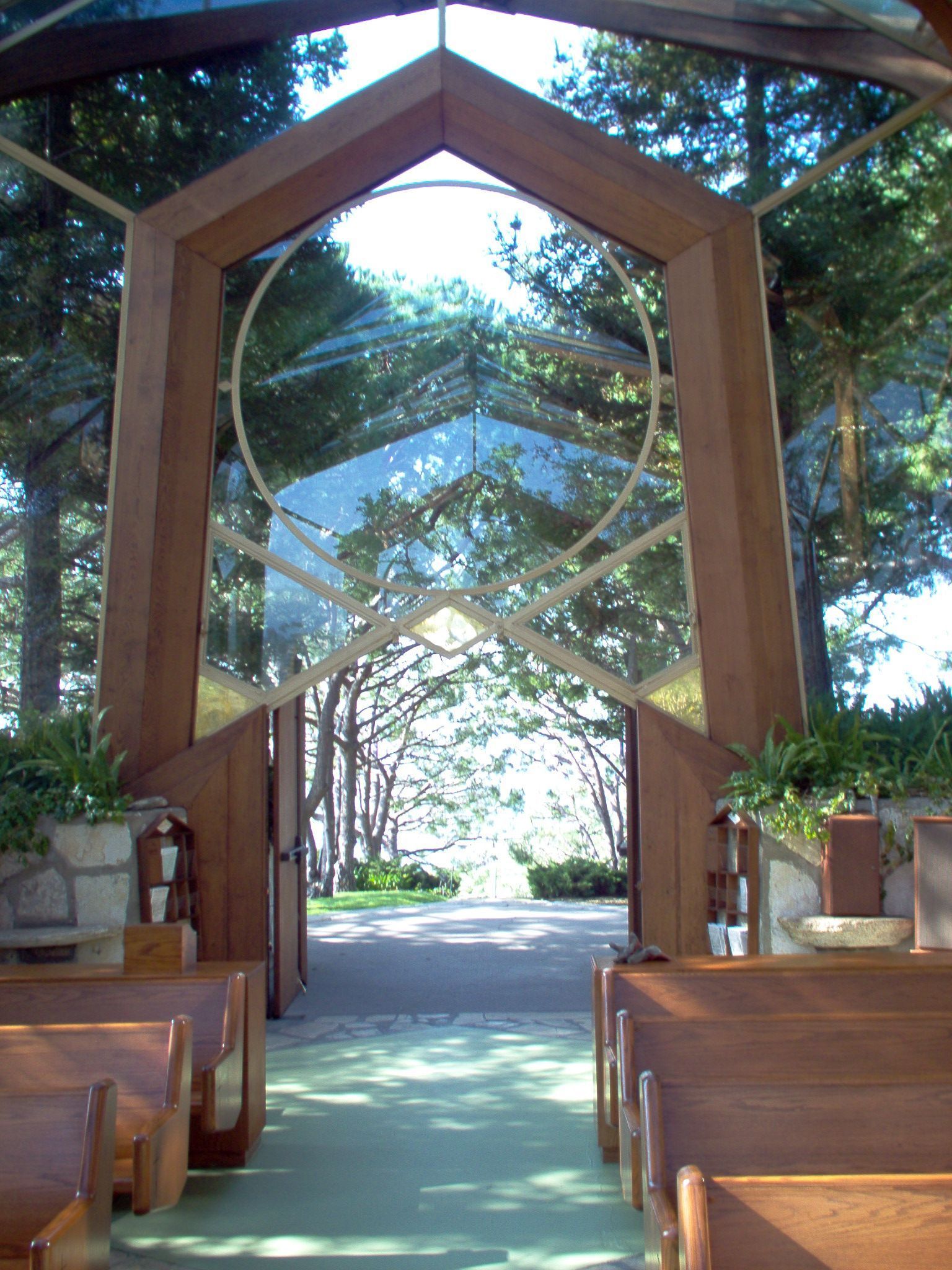 Wooden outdoor wedding aisle beneath a geometric arch, flanked by benches and greenery, leading to bright sunlight. Wooden outdoor wedding aisle beneath a geometric arch, flanked by benches and greenery, leading to bright sunlight.