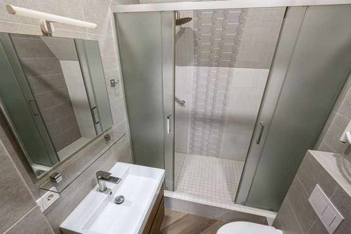 Small bathroom with white sink, mirror, toilet, and glass shower with patterned tile wall.