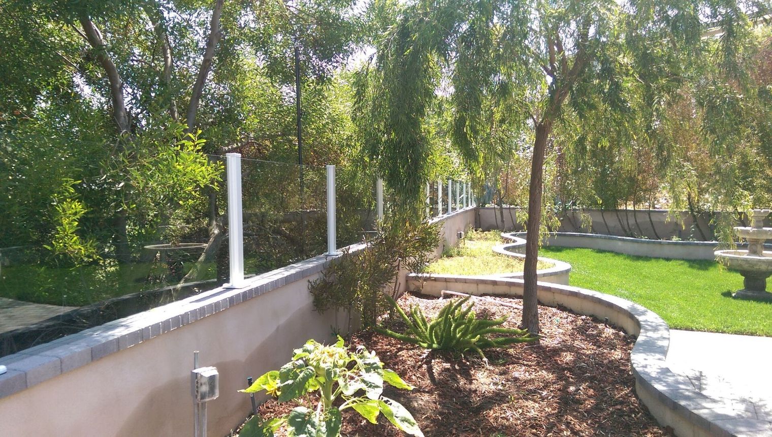 Sunny garden path beside a low wall, with trees, grass, and white railings lining a curved walkway.