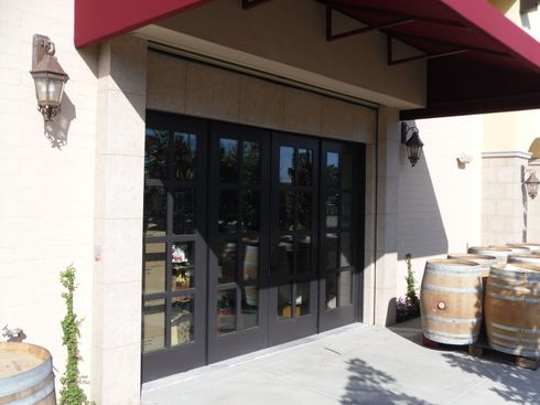 Black glass double doors under a red awning, with wine barrels beside a stucco entrance.