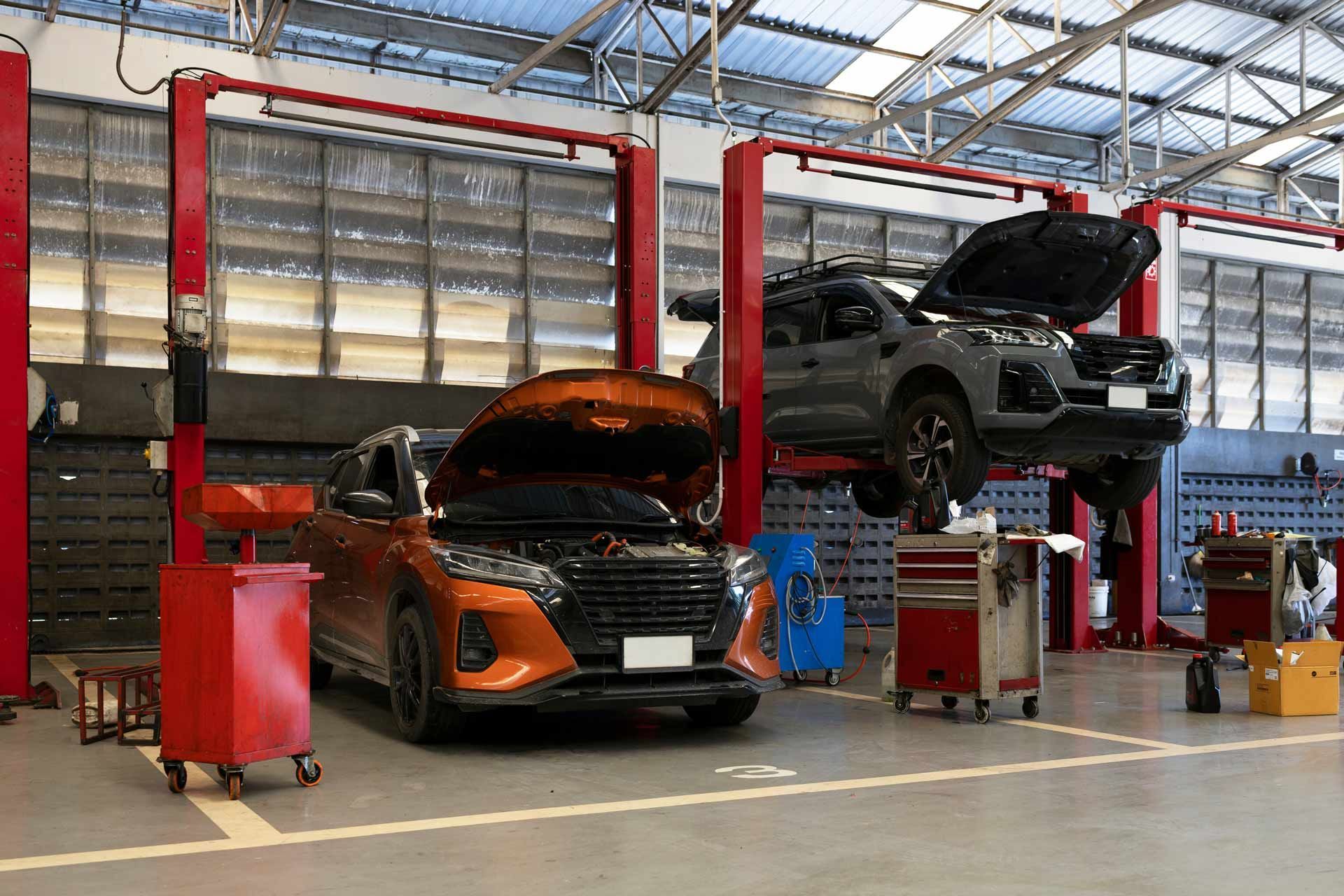 Two cars in an auto repair shop, one orange on the floor and one gray raised on a lift with hood open.