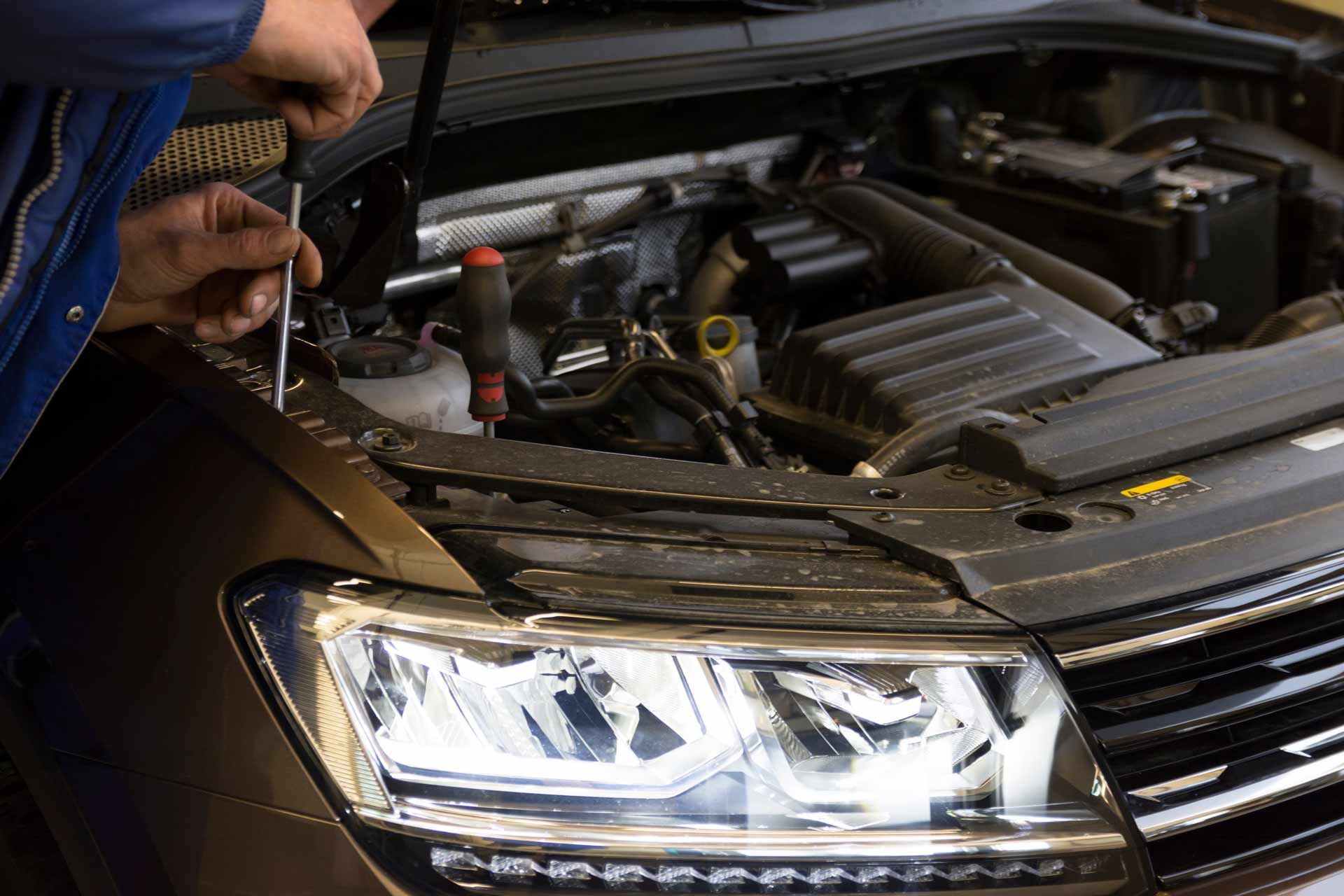 Mechanic inspecting a car engine with the hood open, next to a black sedan's headlight.