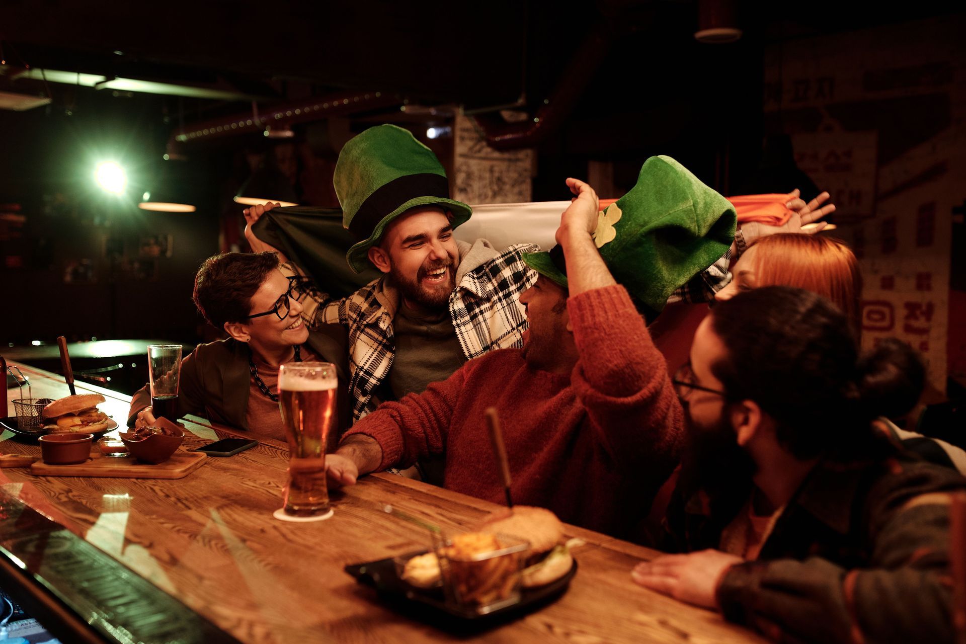 Persone con cappelli verdi che ridono attorno a un tavolo di legno in un bar scarsamente illuminato