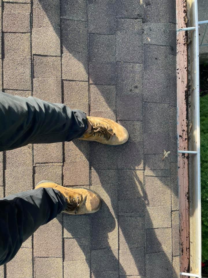 Person’s boots standing on a shingled roof beside a gutter and safety rail.