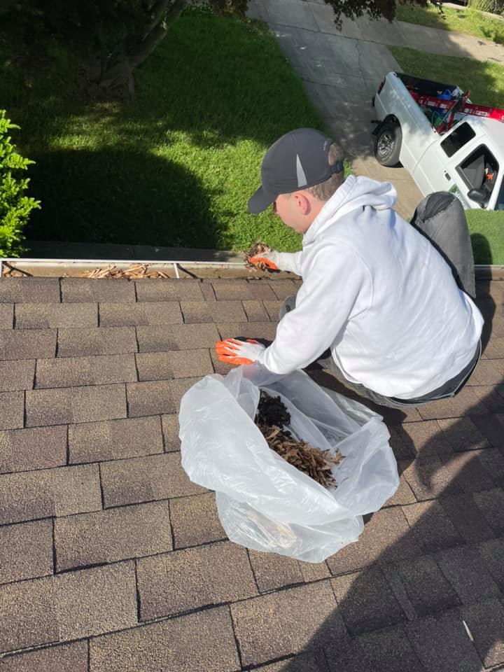 Person in a gray hoodie kneeling on a shingled roof, inspecting debris in a white bag near a gutter.