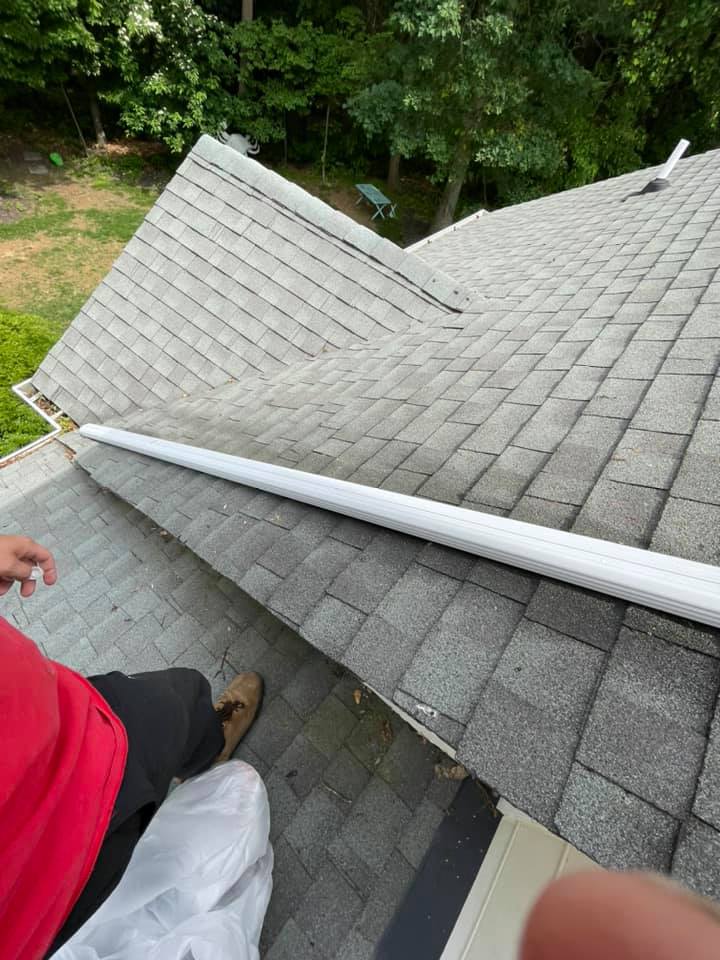 Worker on a gray shingled roof beside a white gutter, with trees and a yard below.
