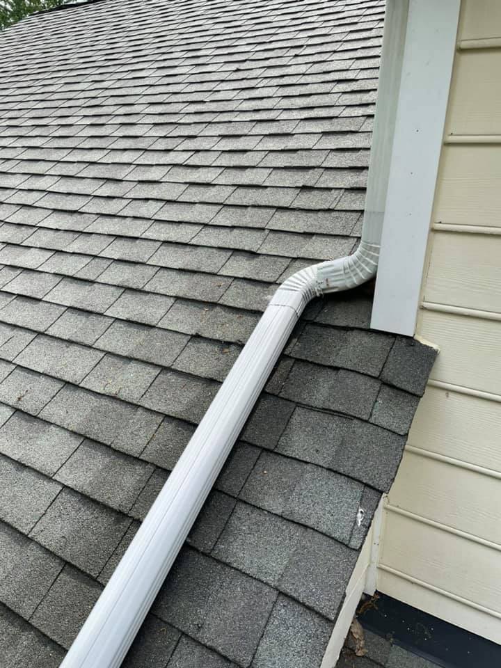 Roof gutter downspout extending over gray shingle roof beside beige siding