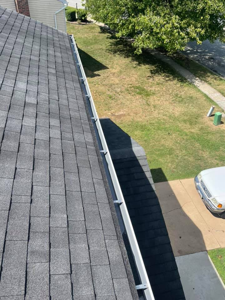 Aerial view of a house roof, driveway, and grassy yard with a parked car and trees.