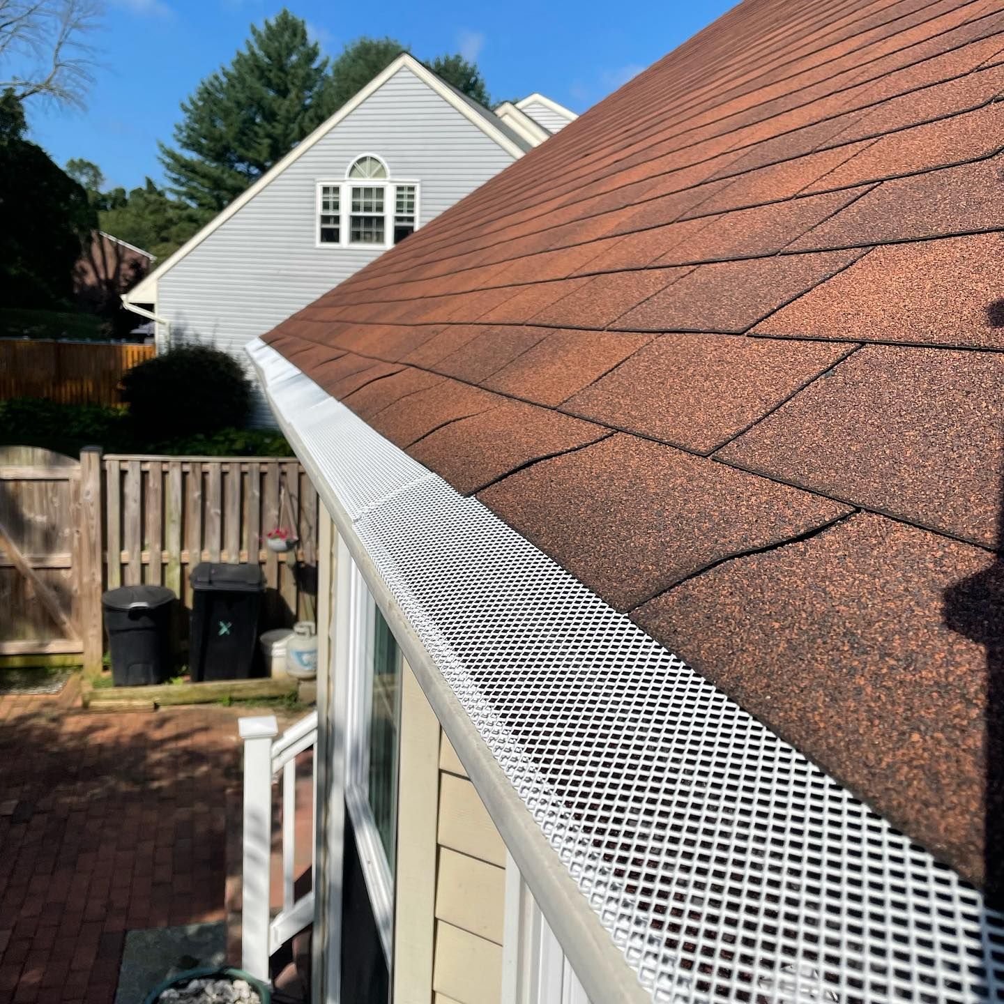 White gutter guard on a red shingle roof beside a beige house and backyard fence