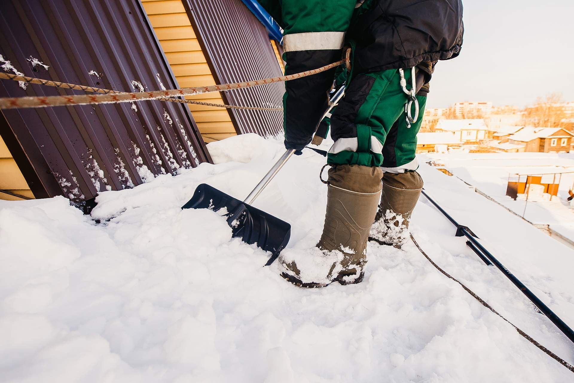 Person shoveling snow beside a corrugated fence in winter, wearing green pants and tan boots