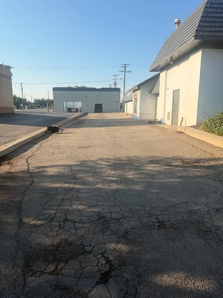 Cracked asphalt driveway between white buildings under clear sky, with a garage in the distance.