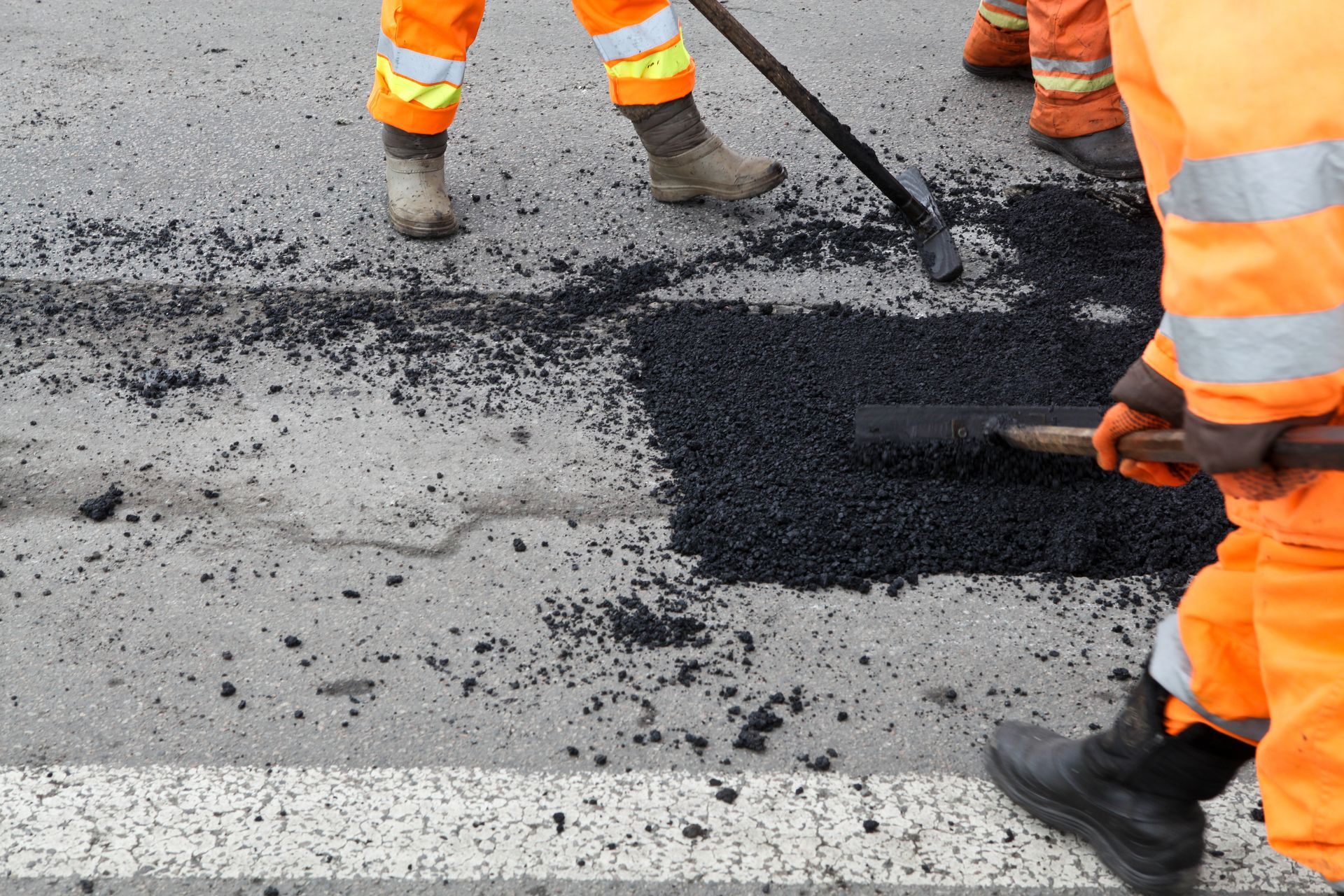Road workers in orange safety gear patching a pothole with black asphalt.