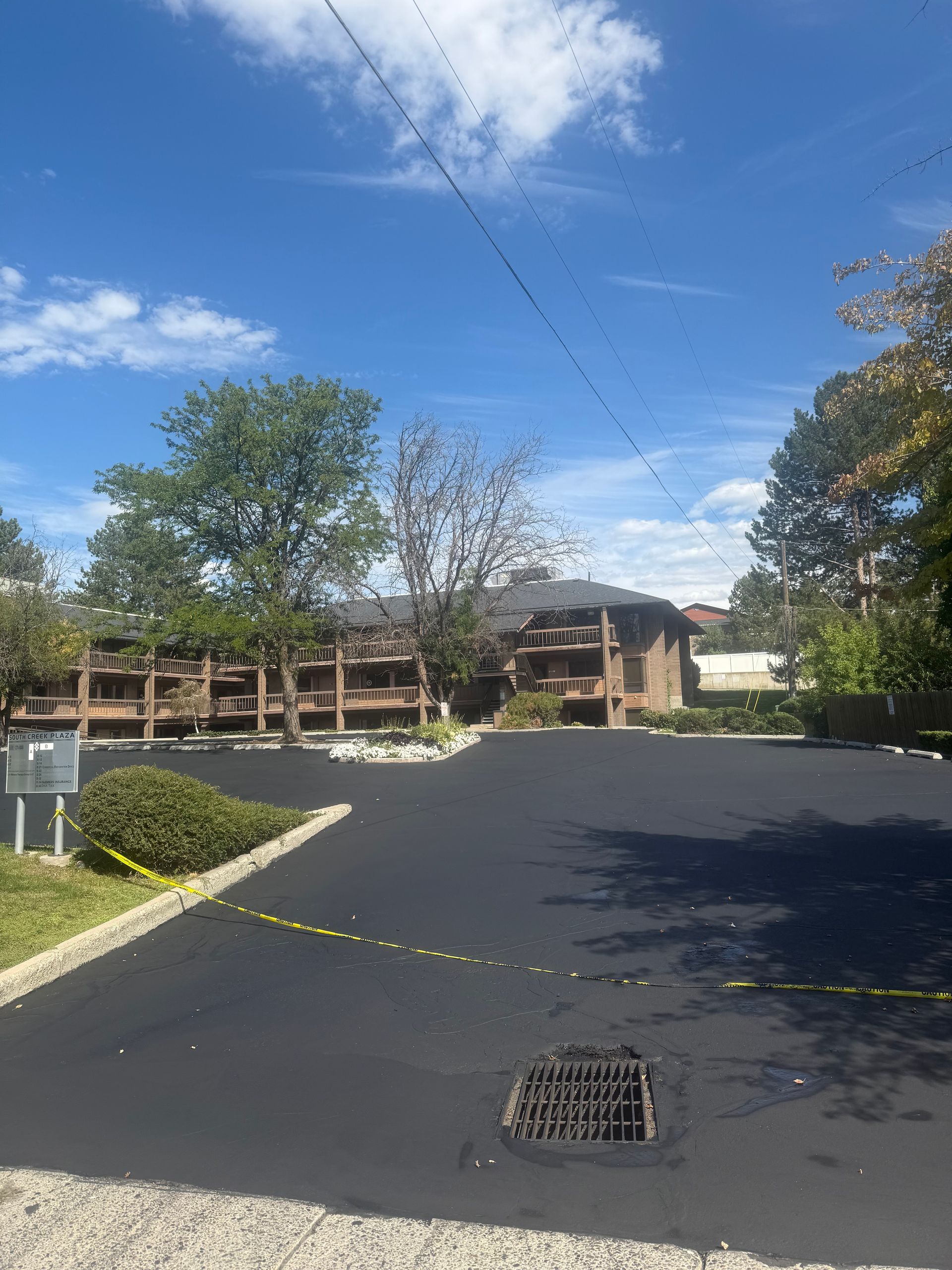 Parking lot with fresh black asphalt, a drain grate, trees, and buildings under a blue sky