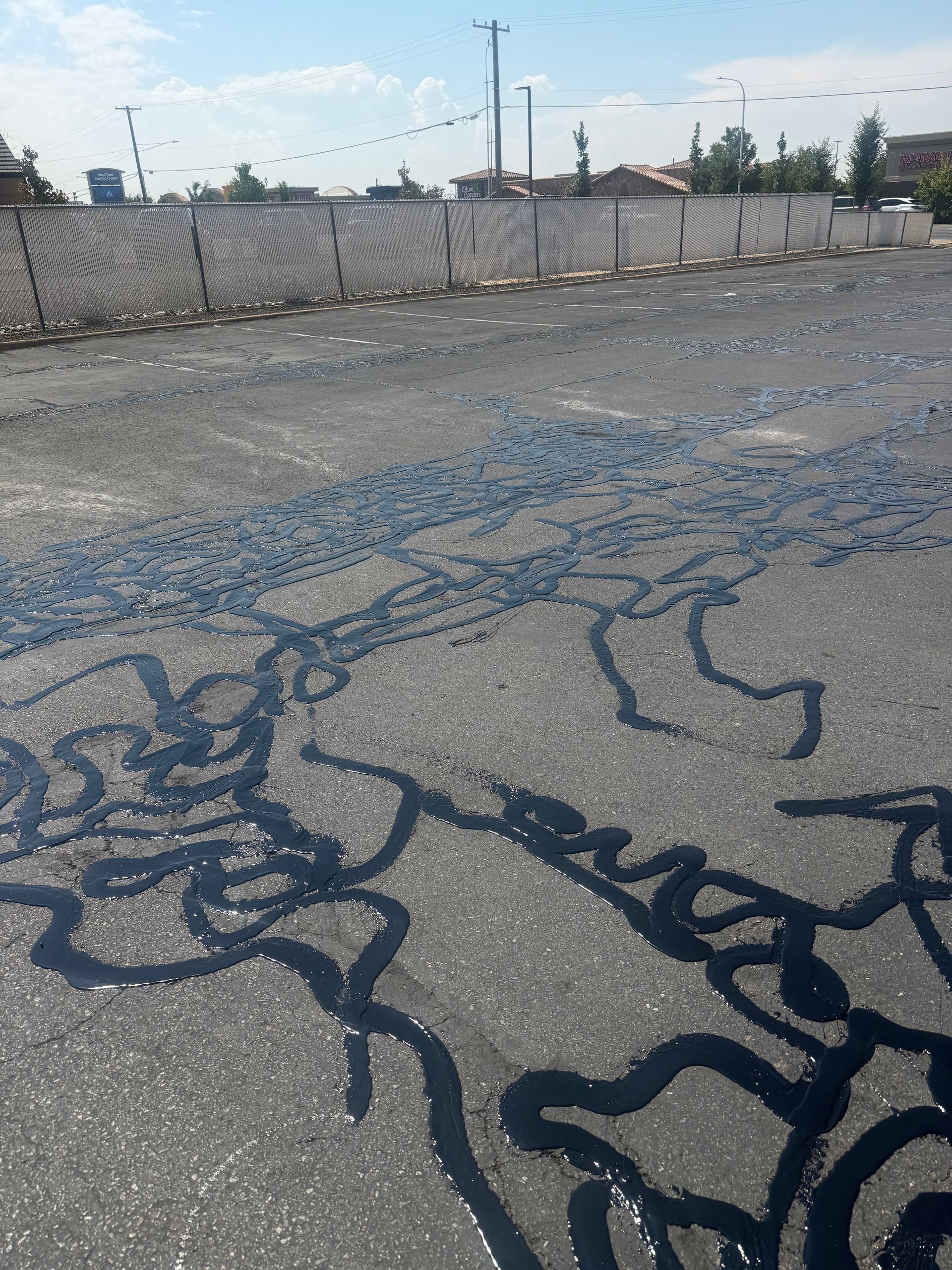 Cracked asphalt lot with dark pavement repairs, chain-link fence, and distant industrial buildings under a clear sky