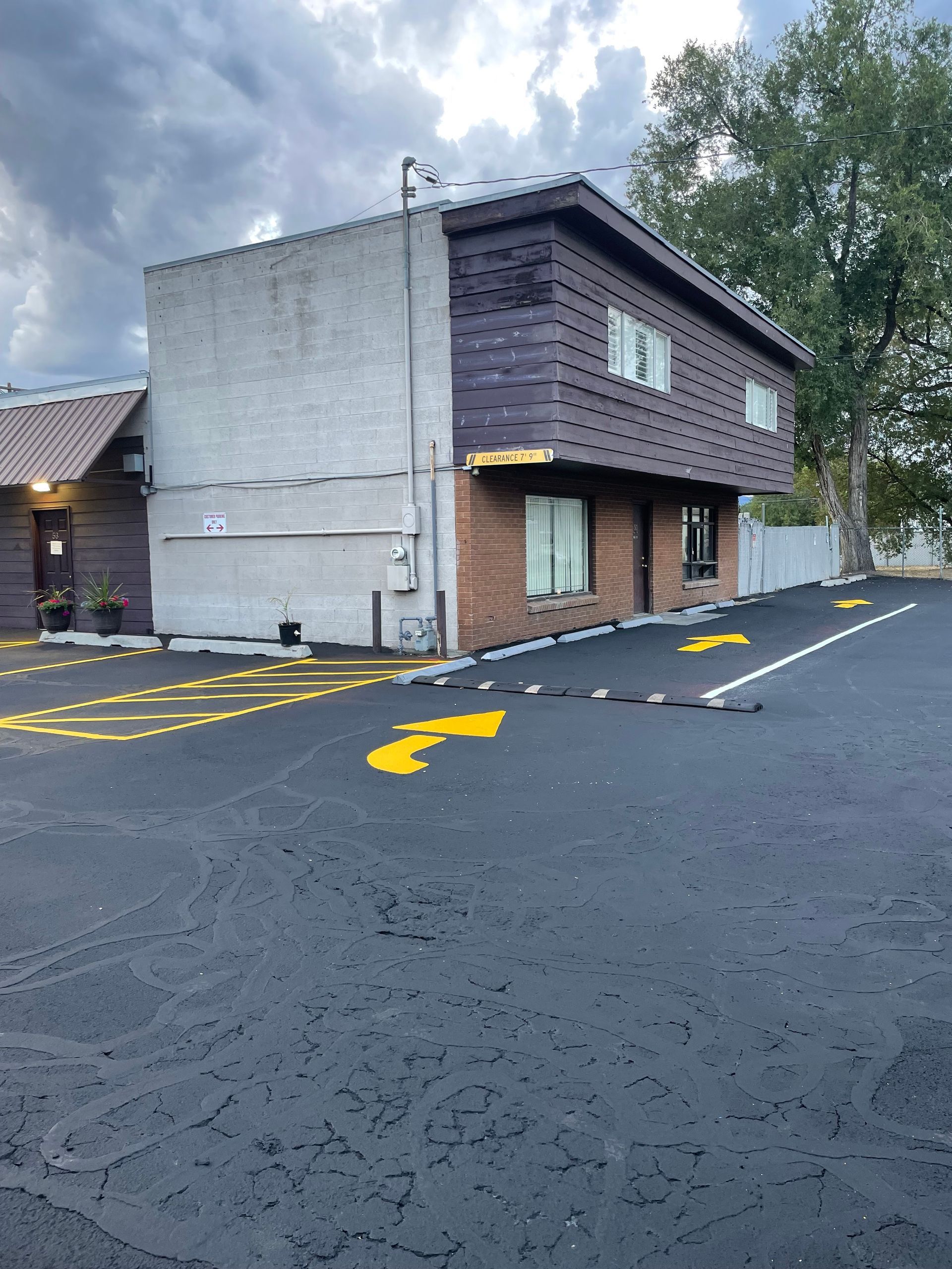 Small two-story brown building beside an empty paved lot with yellow directional arrows and trees nearby.