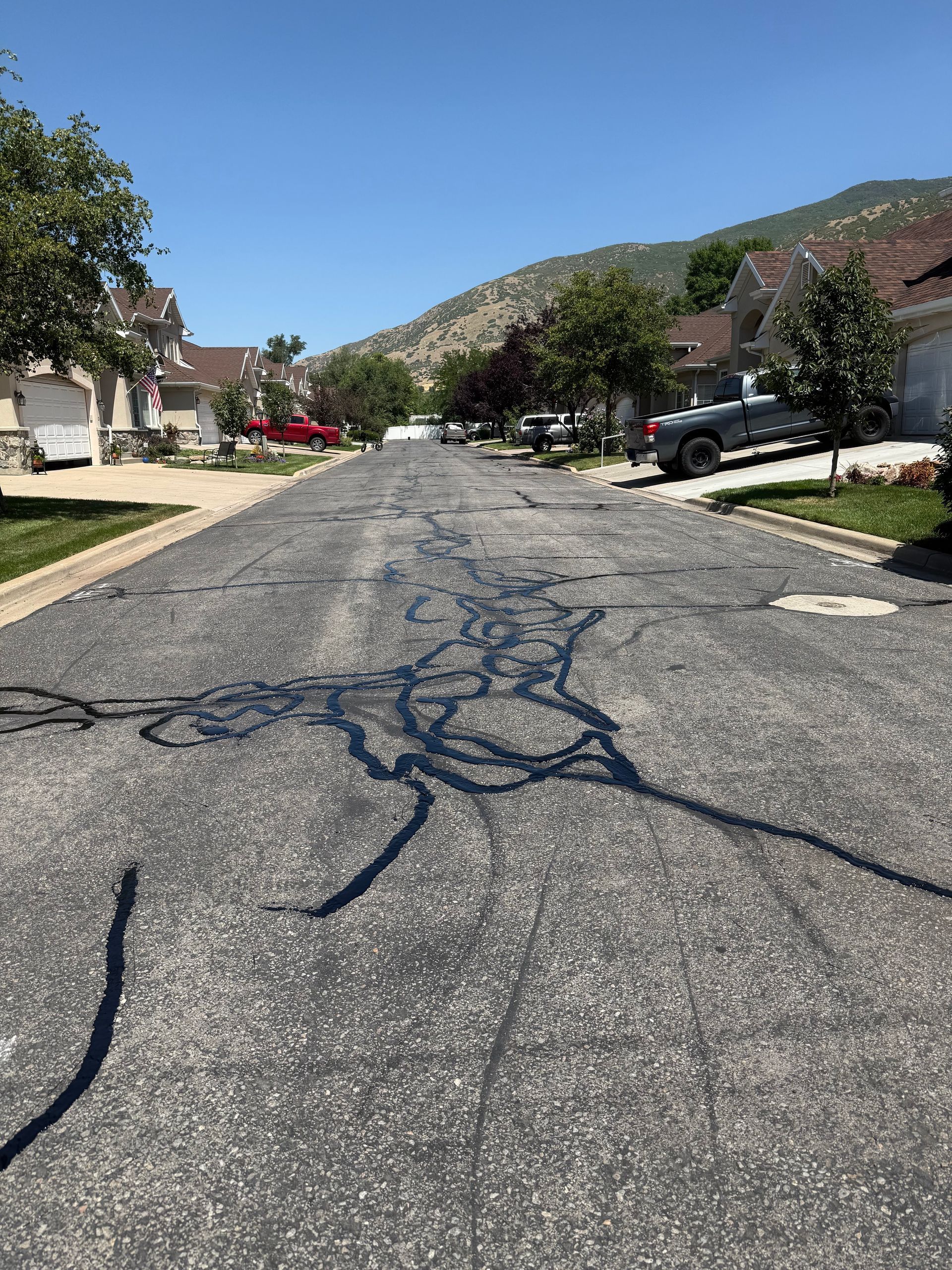 Cracked residential street with parked cars, houses, trees, and hills under a clear sky.