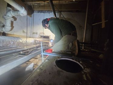 Worker in a dim crawlspace inspecting pipes with a flashlight near a large floor opening