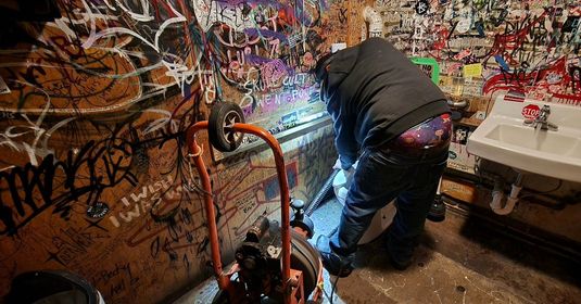 Person in a dark jacket leaning over a cluttered graffiti-covered table in a small, colorful room