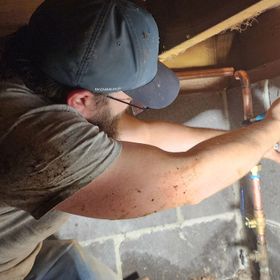 Man in a cap and glasses working on copper pipes in a crawlspace, reaching with tools.