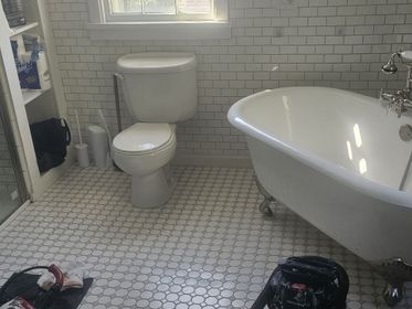 Small white-tiled bathroom with a toilet, freestanding tub, window, and laundry baskets on the floor