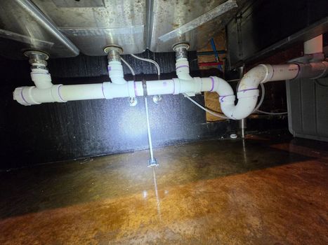 Leaking white PVC pipes under a sink, with water spraying onto the floor.