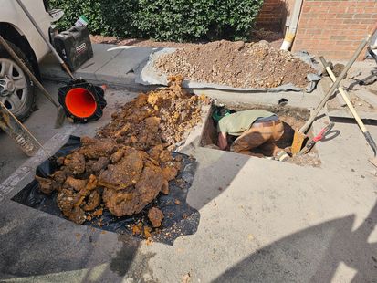 Construction trench beside a sidewalk with excavated dirt, a bucket, and exposed underground utility pipe.
