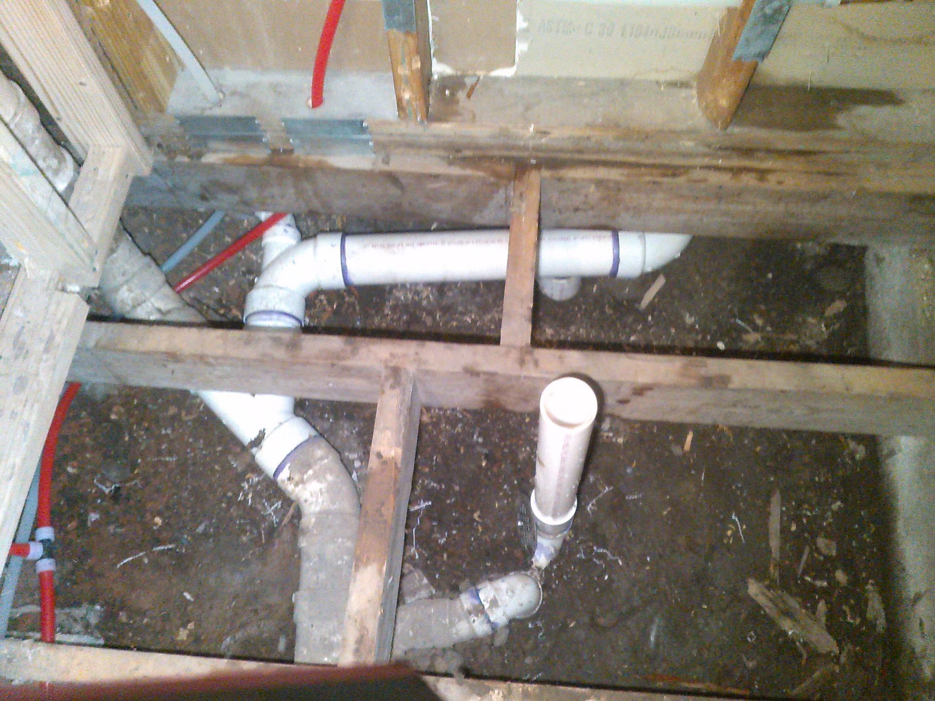 Exposed white PVC pipes and wooden framing beneath a floor opening, with dirt and debris below.