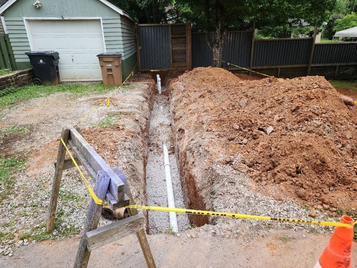 Open trench beside gravel driveway with orange soil piles, caution tape, and a garage in the background